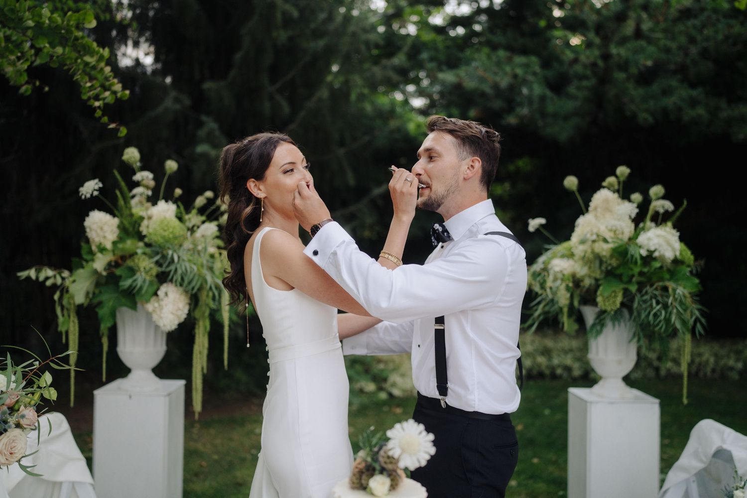 Couple playfully feeding each other cake at outdoor wedding ceremony.