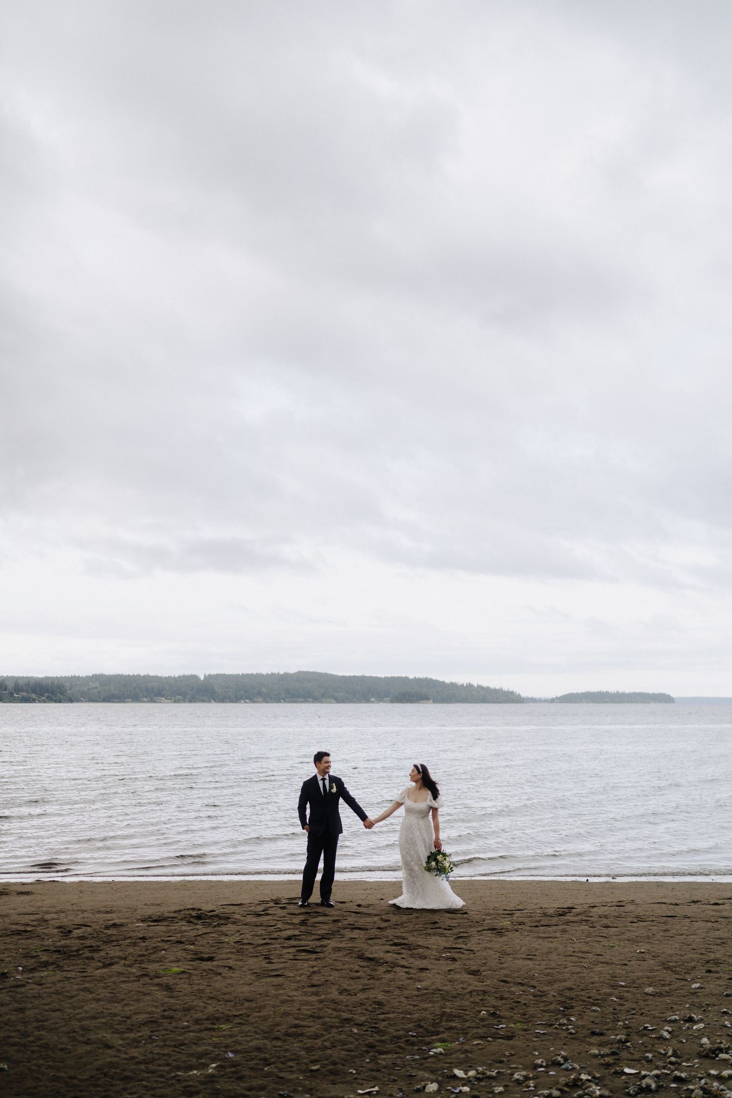 A couple in wedding attire walking along a misty beach shoreline.