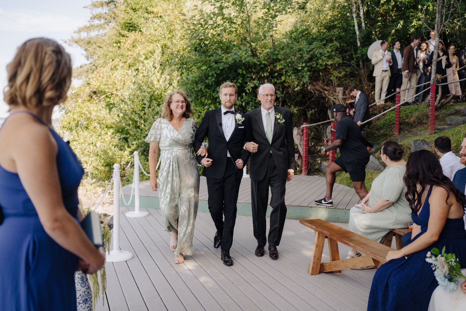 Wedding guests gather outdoors on a wooden deck surrounded by greenery during a ceremony.