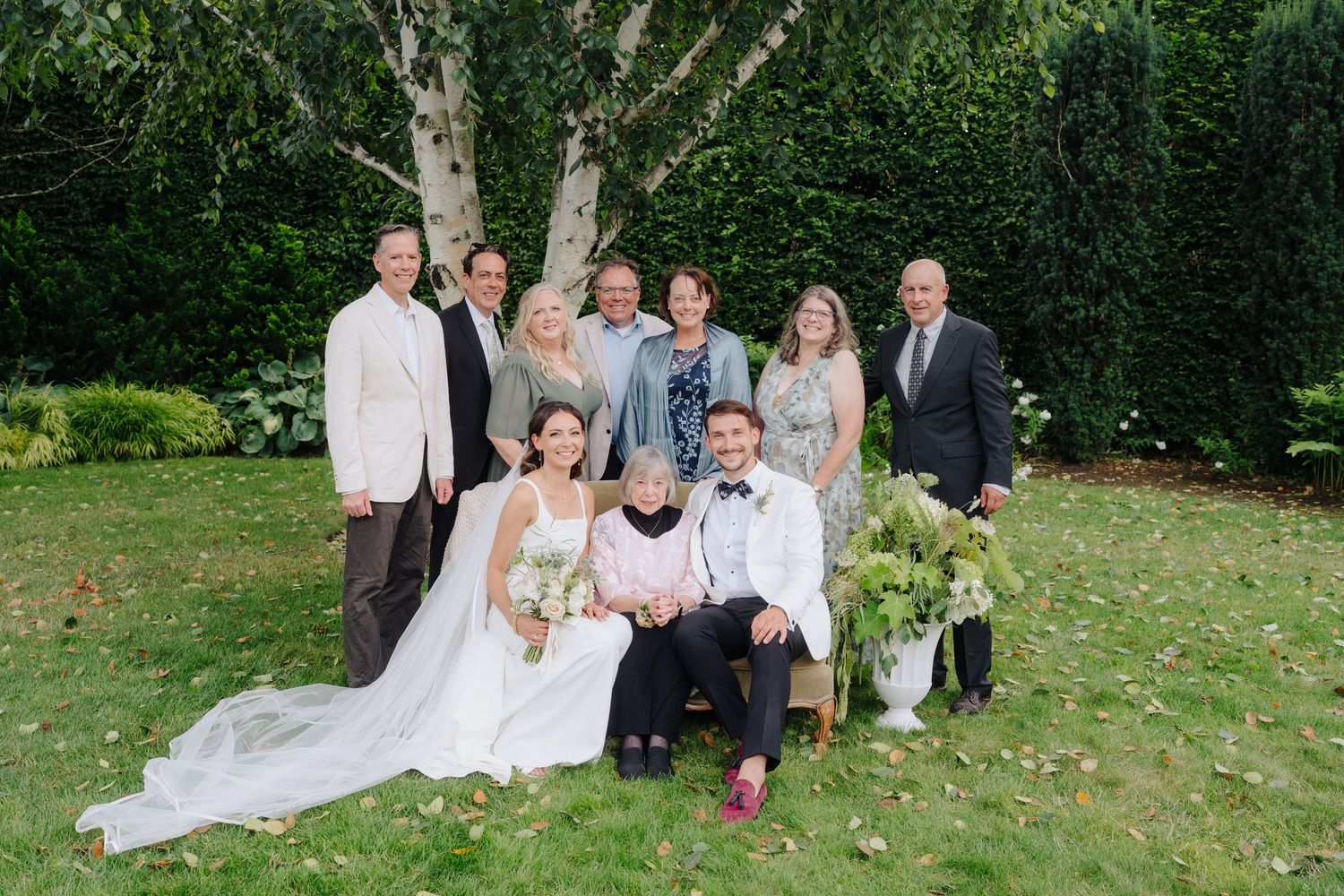 Wedding party poses together on green lawn with birch trees and hedges in background.