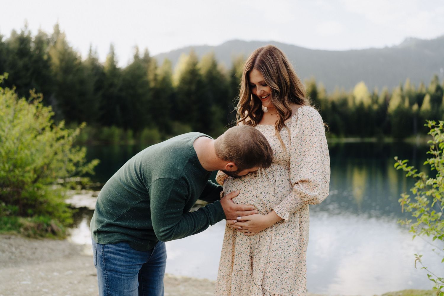 Couple embracing by a serene mountain lake with evergreen trees and mountains in the background.