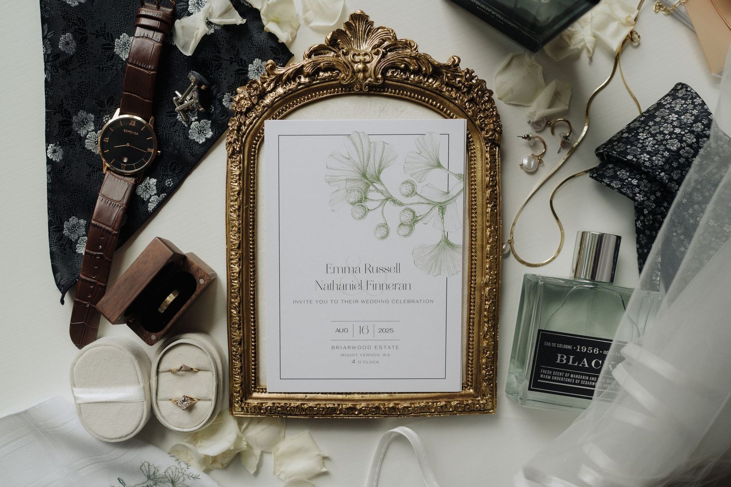 Elegant wedding invitation in ornate gold frame with beauty products and accessories on white fabric.