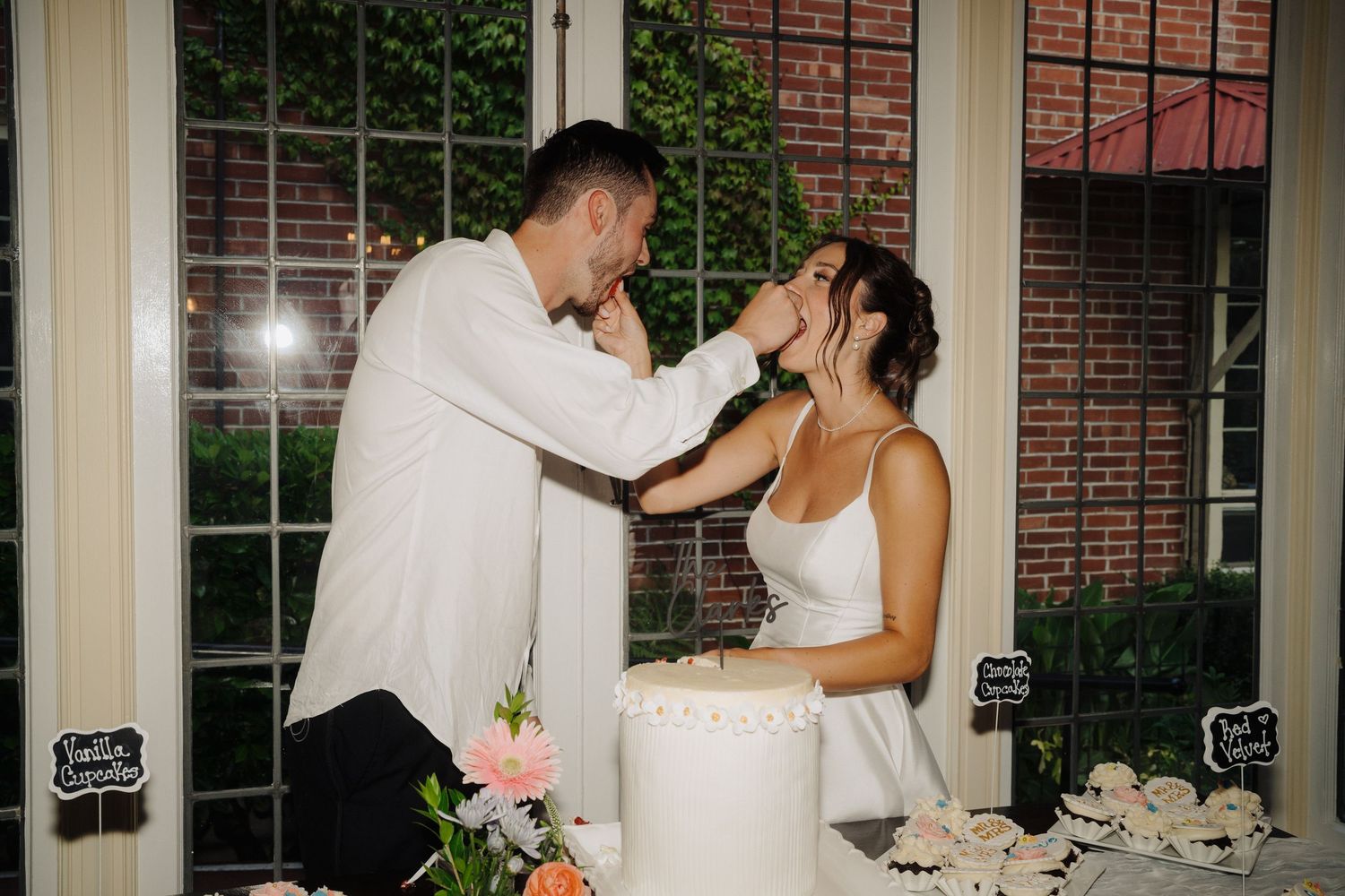 Wedding cake cutting moment captured indoors with floral decorations and vintage windows.