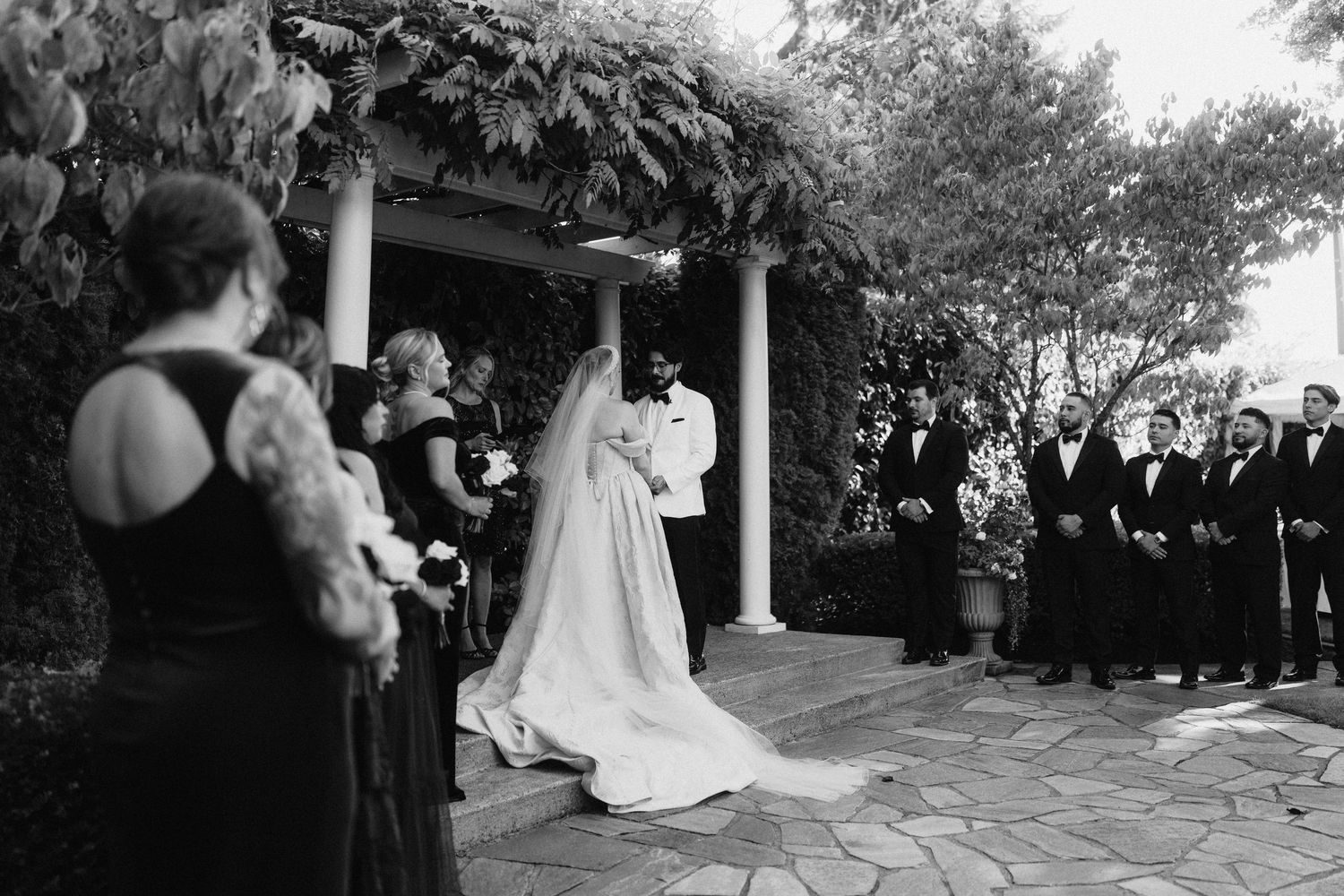 Black and white photo of wedding guests standing under a columned porch during an outdoor ceremony.
