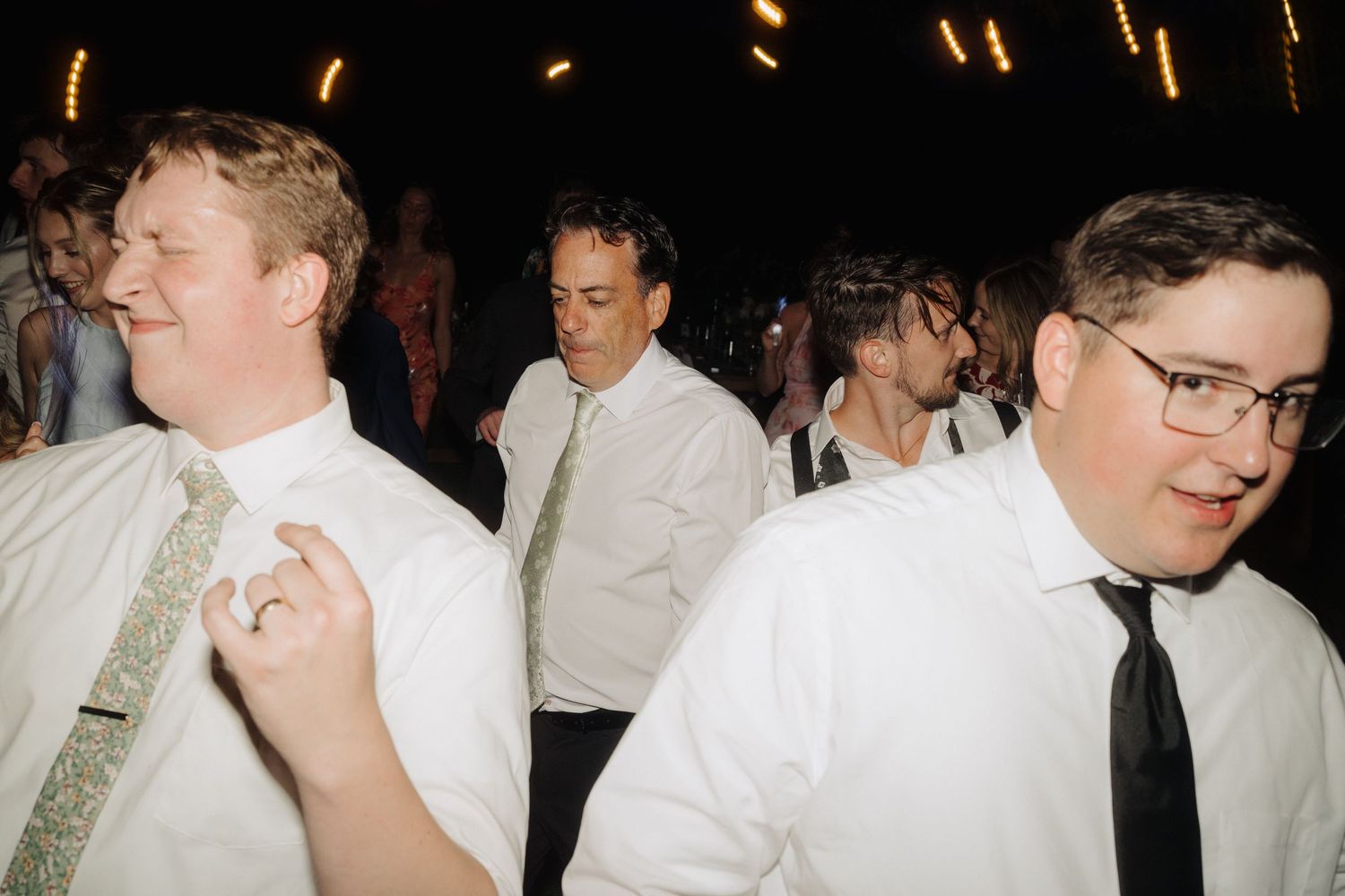 Wedding guests in formal attire dance and socialize at a dimly lit reception.
