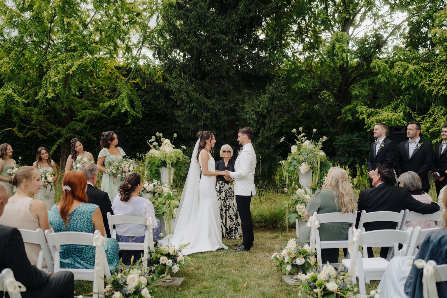 Outdoor wedding ceremony setup with white chairs and floral arrangements in a garden setting.