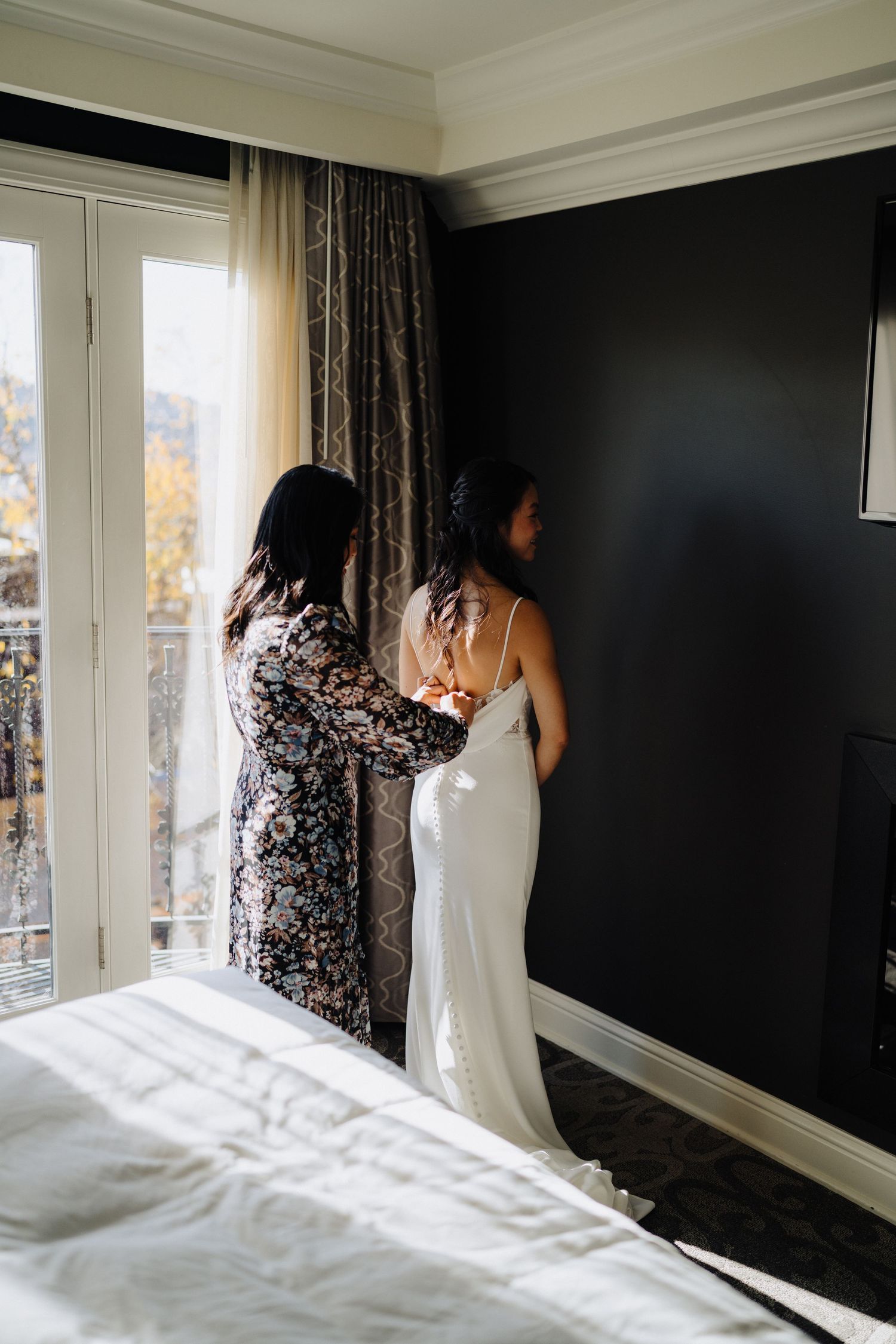 A white wedding dress being adjusted in a sunlit room with dark walls and elegant curtains.