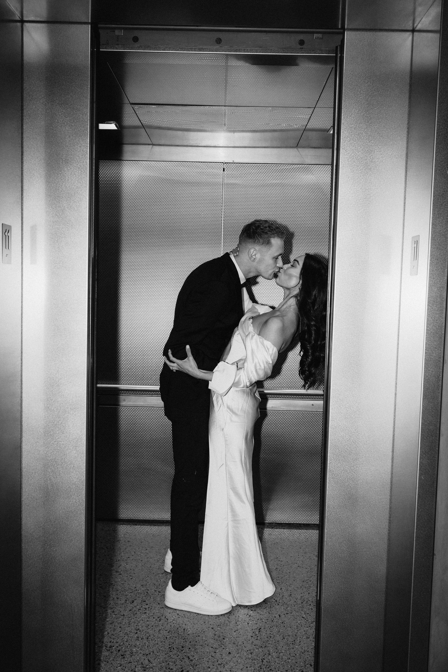 A romantic black and white photo of a couple sharing a kiss in an elevator wearing formal attire.