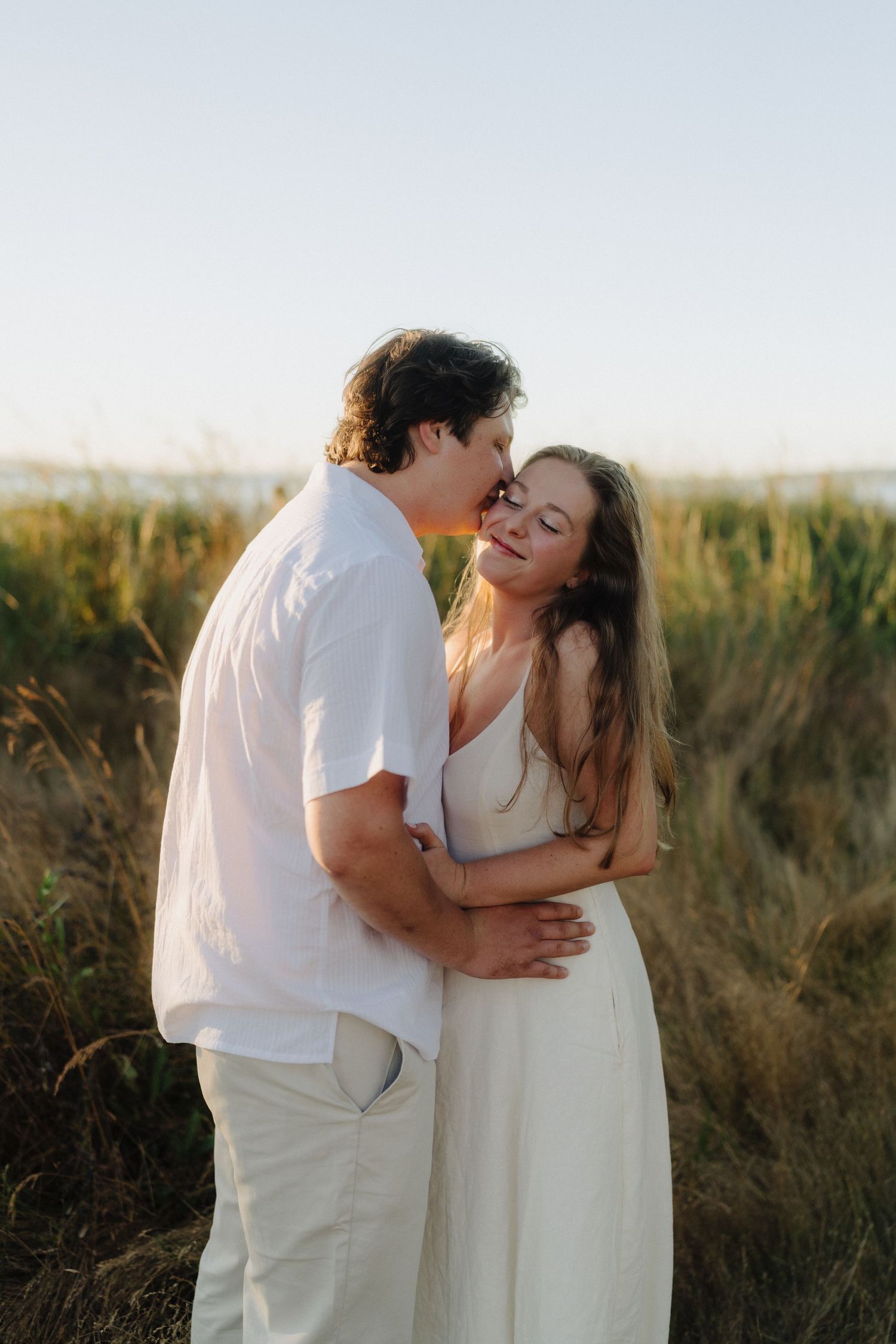 A romantic couple in white outfits share intimate moments in a field of tall grass during golden hour.