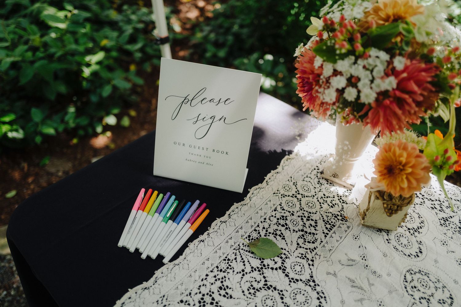 Wedding guest book and colorful pens displayed with floral centerpiece on lace-covered table.
