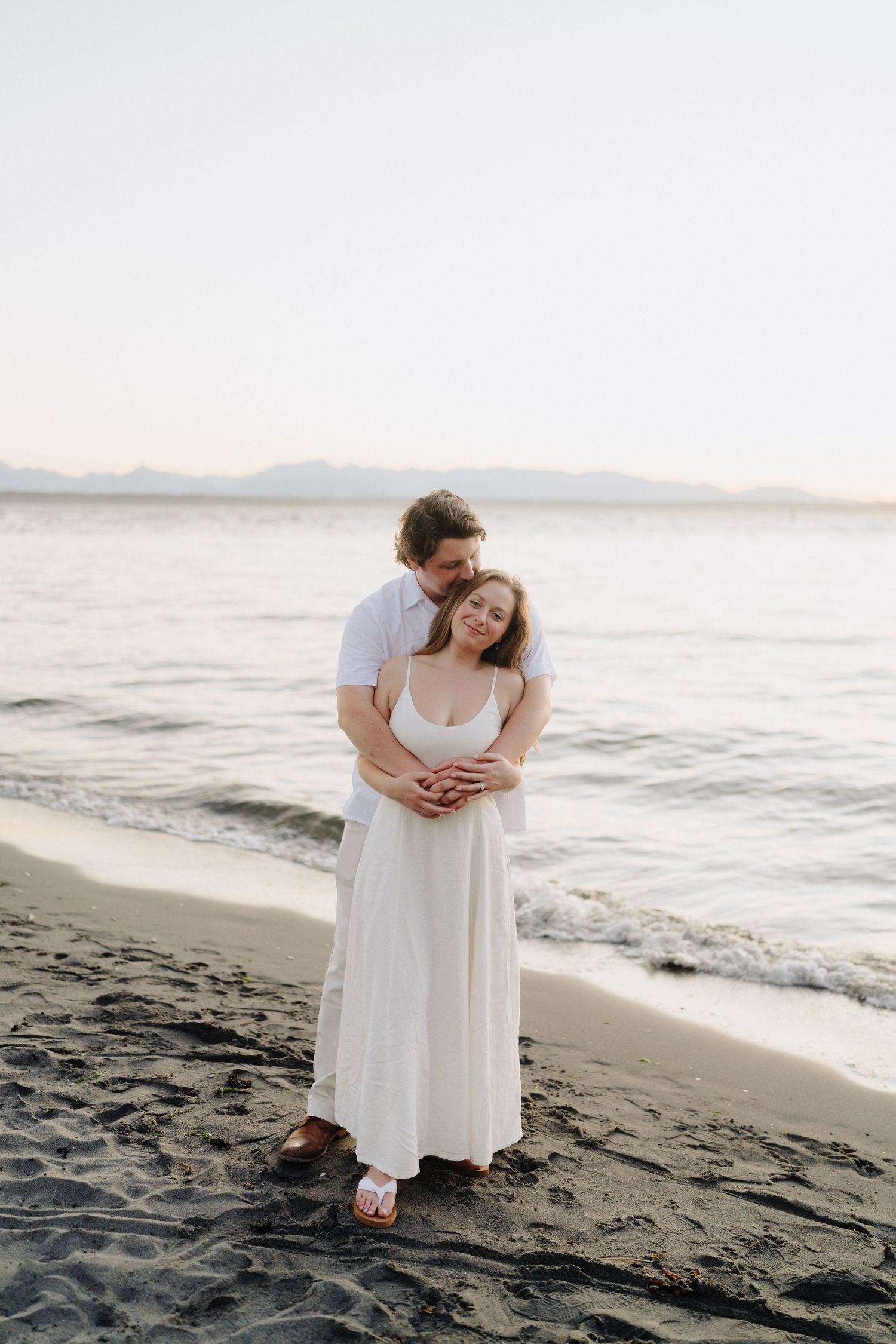 A couple in white outfits shares intimate moments on a dark sandy beach at sunset.
