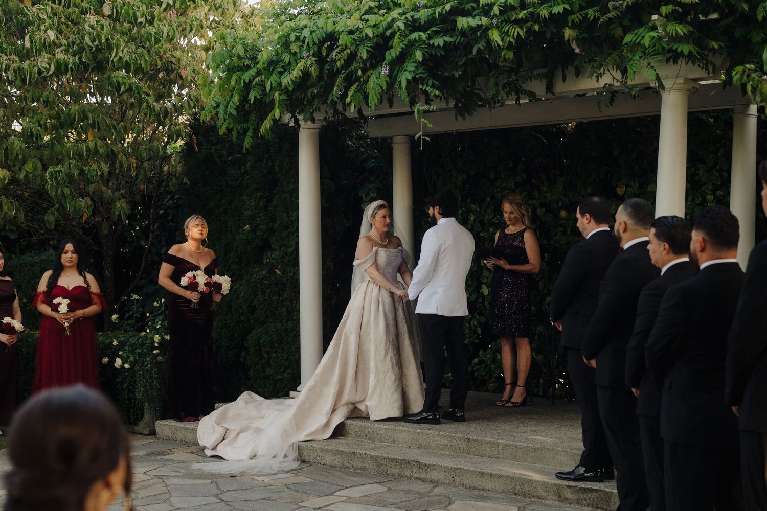 Wedding guests gather on a lush garden patio surrounded by greenery during an outdoor ceremony.