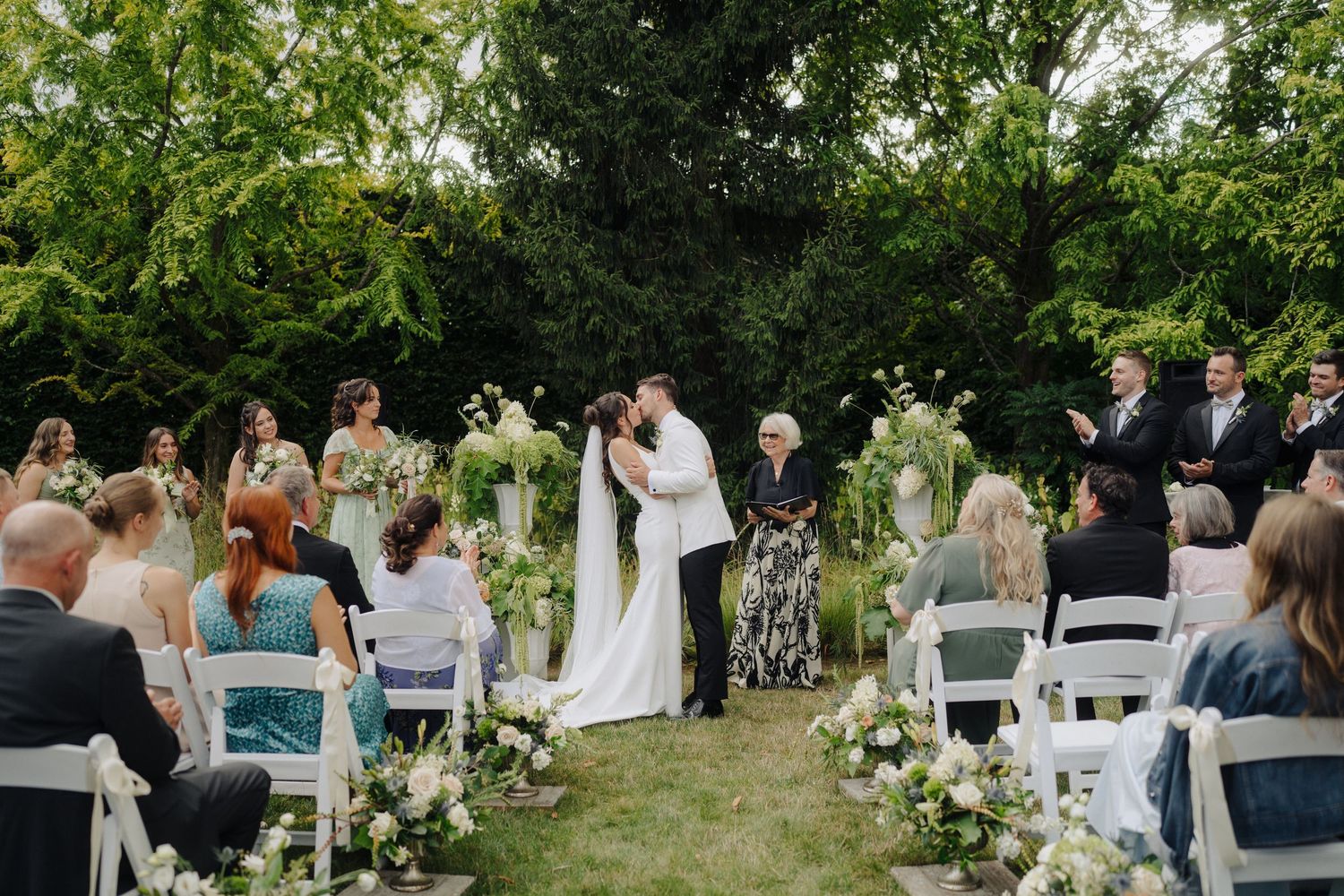 Wedding guests seated in white chairs during an outdoor garden ceremony.
