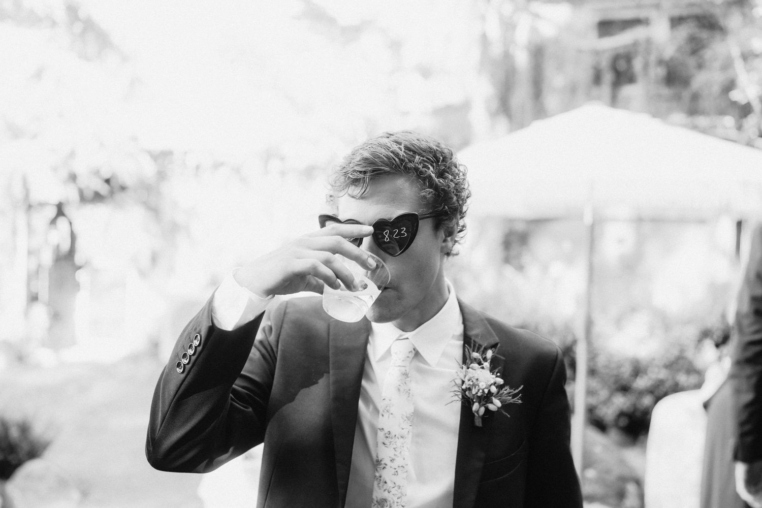 Black and white portrait of suited guest enjoying drink at outdoor wedding reception.
