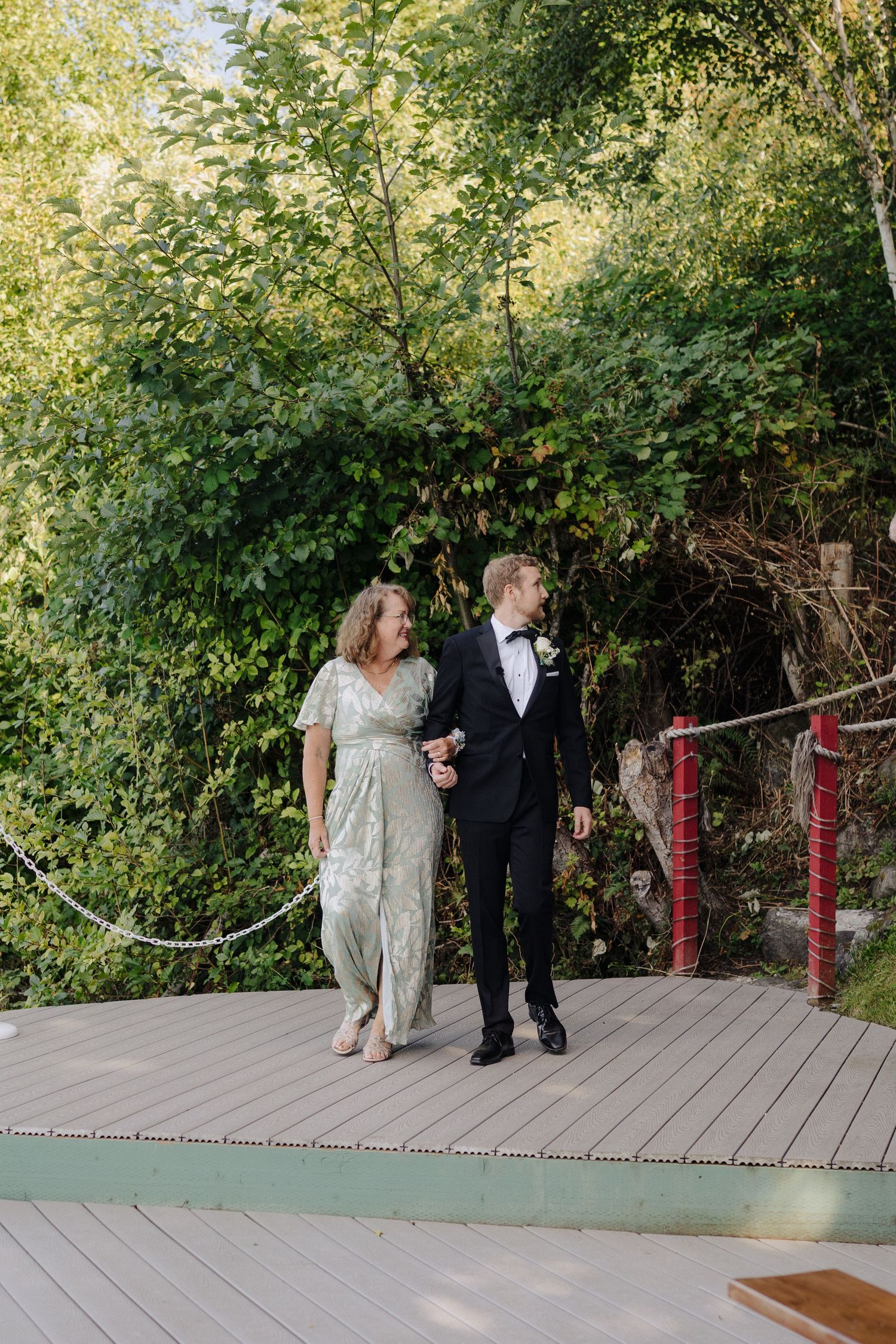 A couple walks together on a wooden deck surrounded by lush green foliage and red posts.