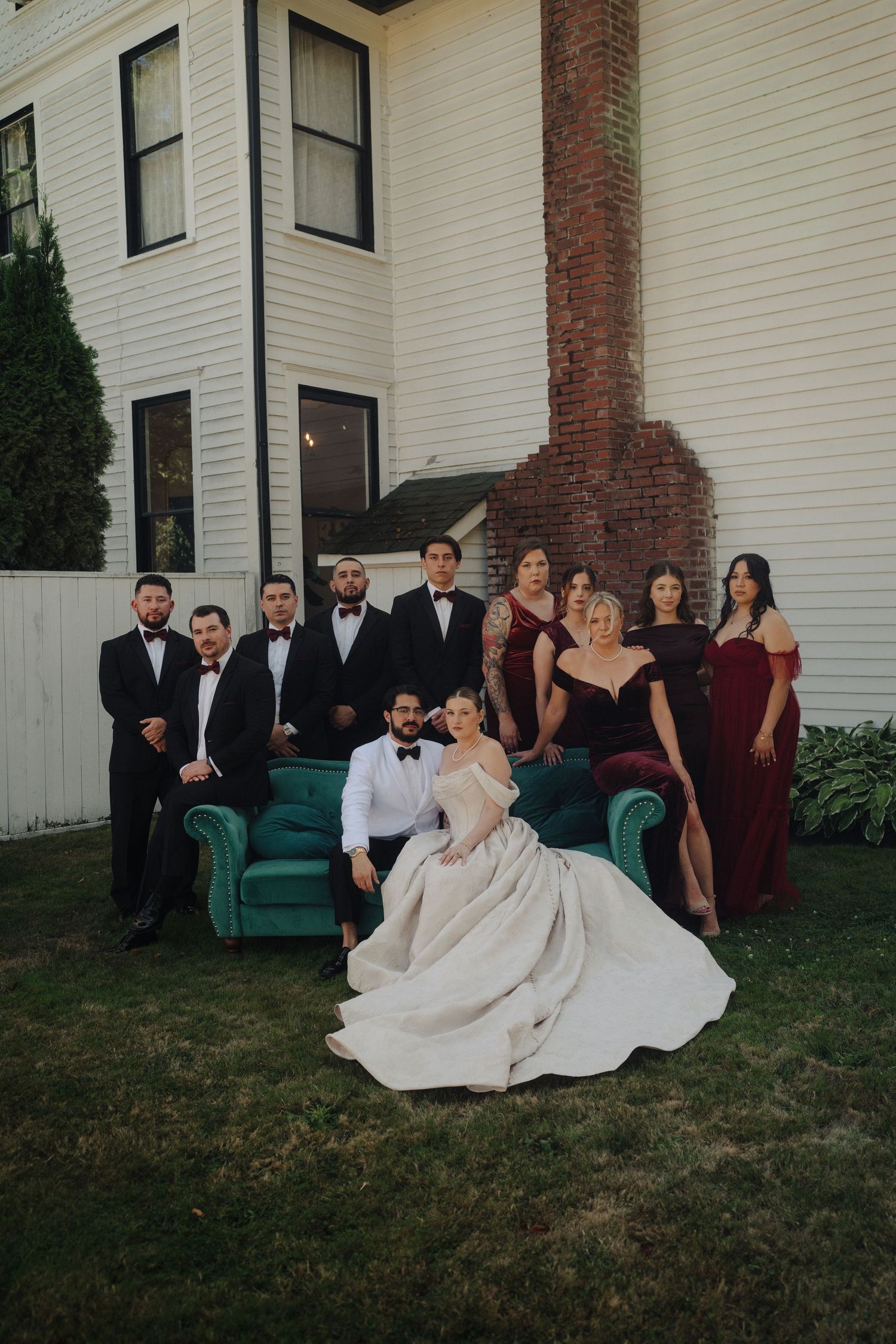 Wedding party poses on vintage teal couch outdoors against white house with brick columns.