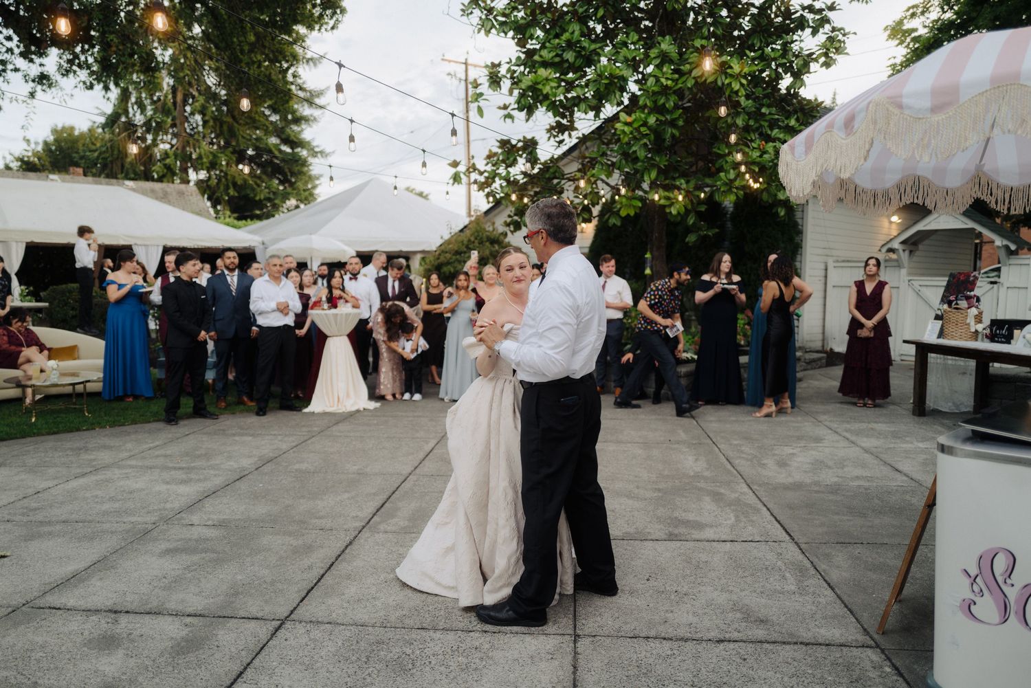 Wedding reception guests dancing on outdoor patio under string lights and tent.