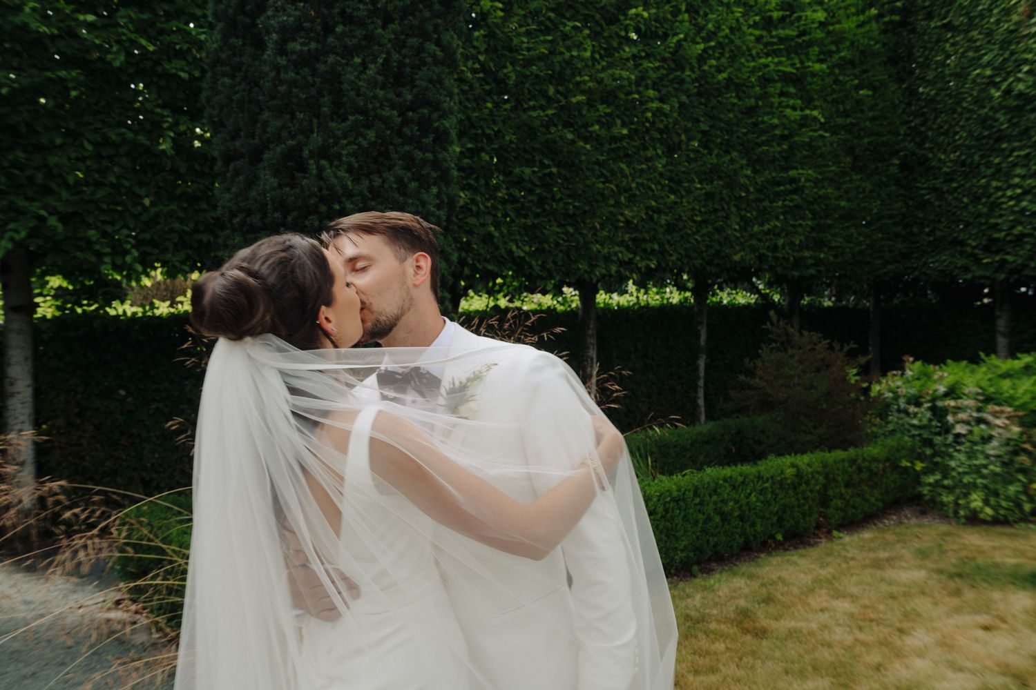 Romantic wedding couple embracing on a garden path with lush foliage backdrop.