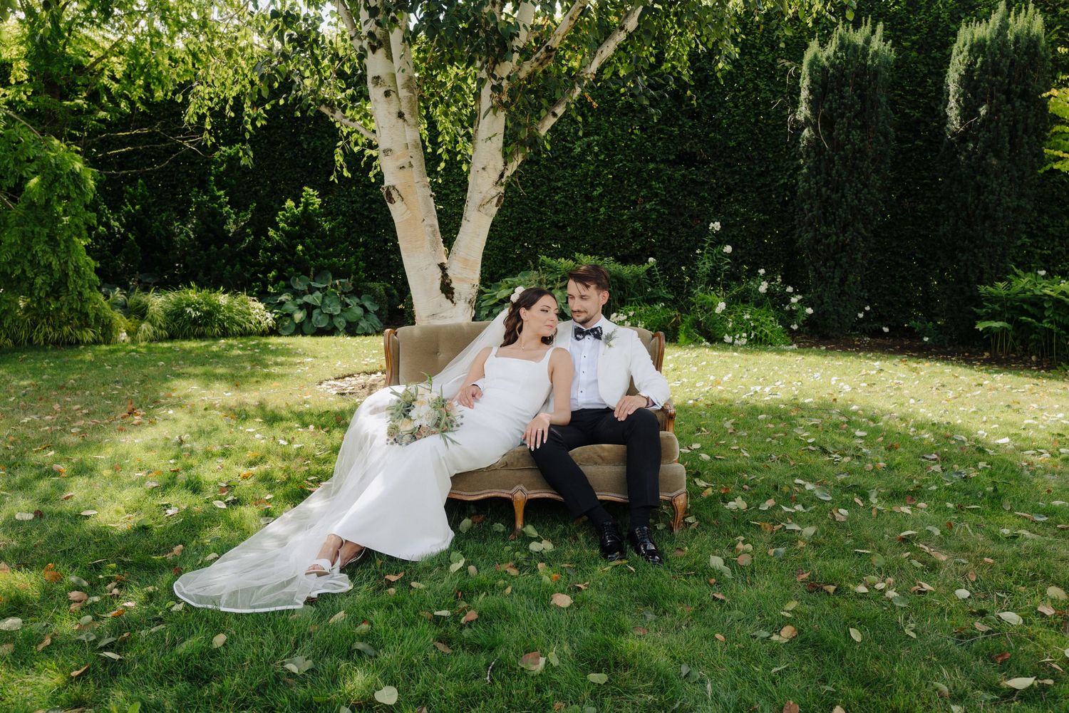 A couple in wedding attire embraces on a garden bench surrounded by greenery and natural light.