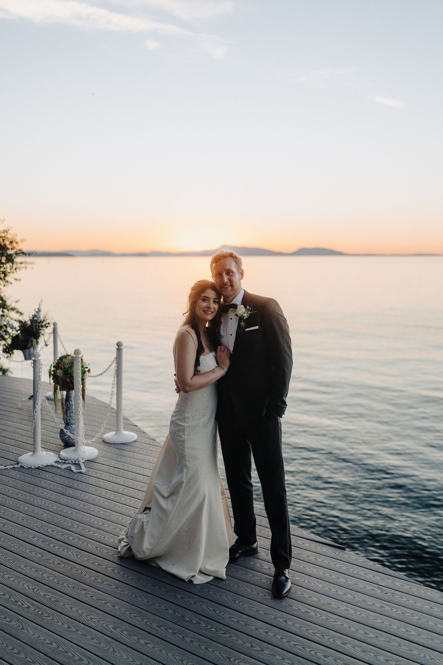A couple embraces on a waterfront dock during sunset with mountains in the background.