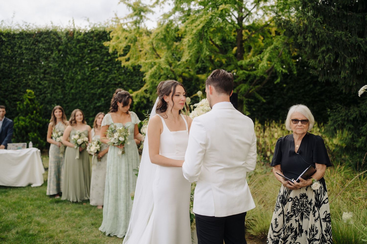 Wedding ceremony guests seated in white chairs on a grassy lawn surrounded by trees and greenery.