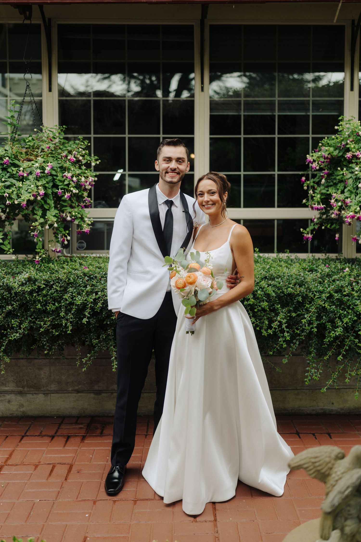 A wedding couple poses in front of a window with pink flowering bushes and brick pavers.