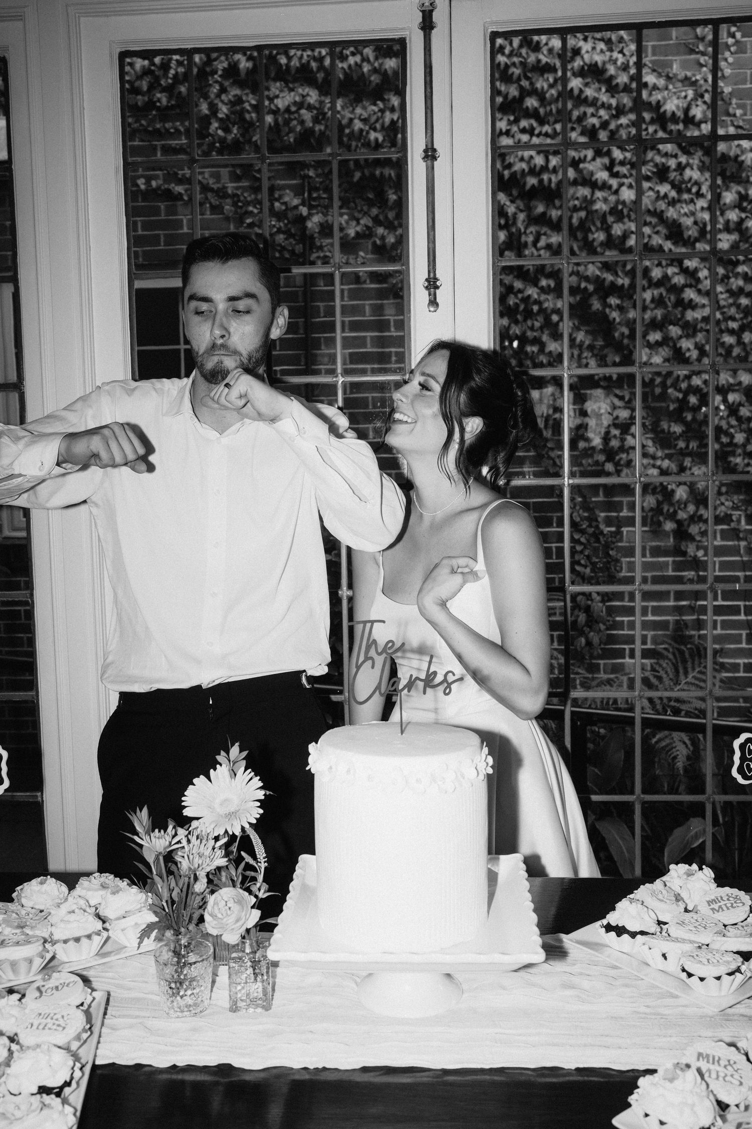 Black and white photo of couple cutting their wedding cake surrounded by floral decorations.