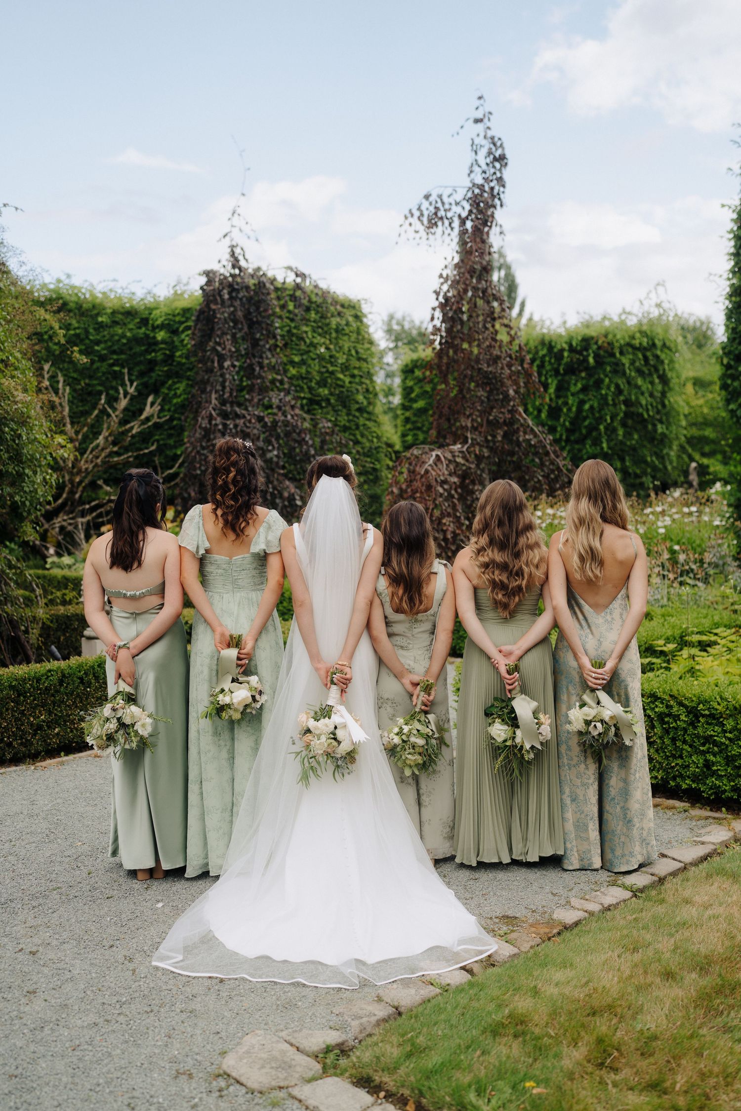 Bridesmaids in sage green dresses stand with bride in white gown holding bouquets in a garden setting.