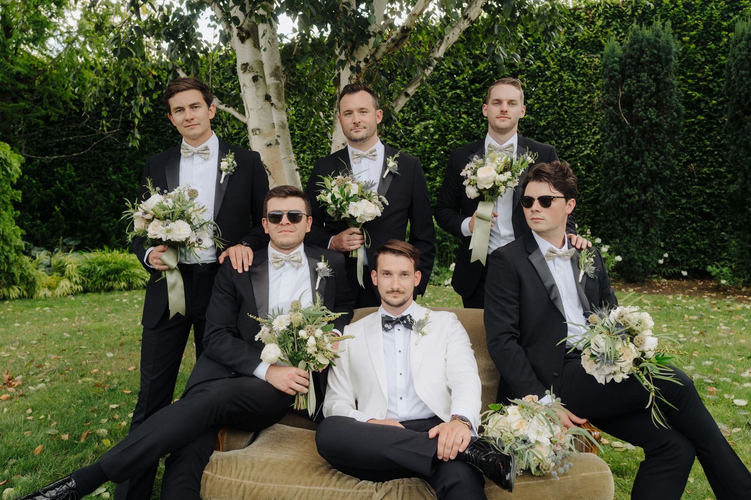 Wedding party members in black suits and bow ties posing together outdoors with greenery backdrop.