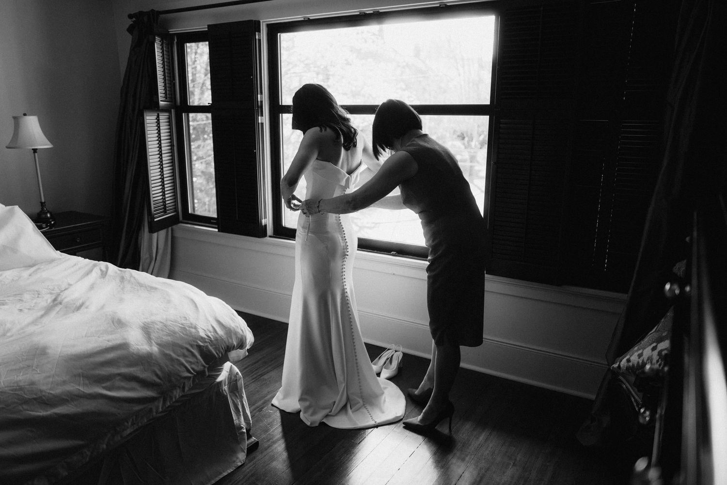 A black and white photograph of two people in a bedroom, one adjusting the other's elegant white wedding gown.