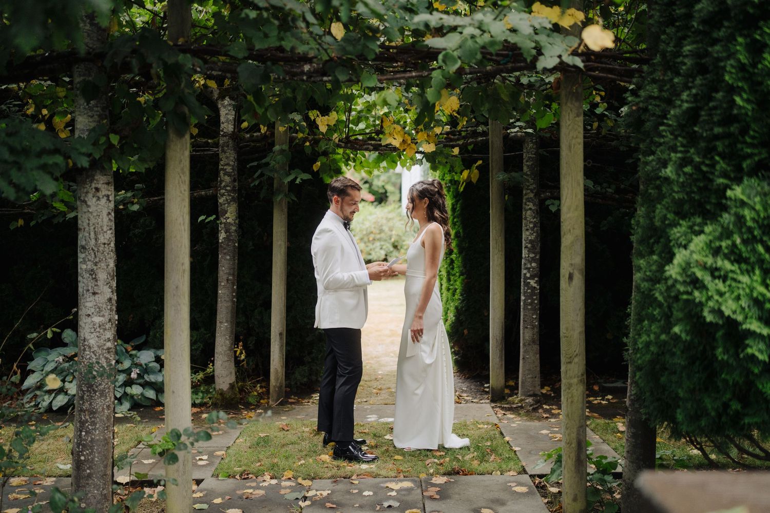 Wedding couple walks through a lush garden path lined with tall trees and greenery.