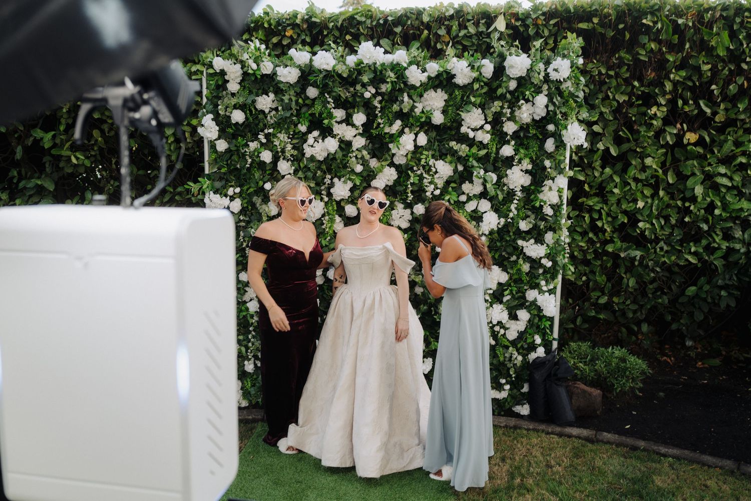 Wedding party posing in front of a lush floral wall backdrop with white roses and greenery.