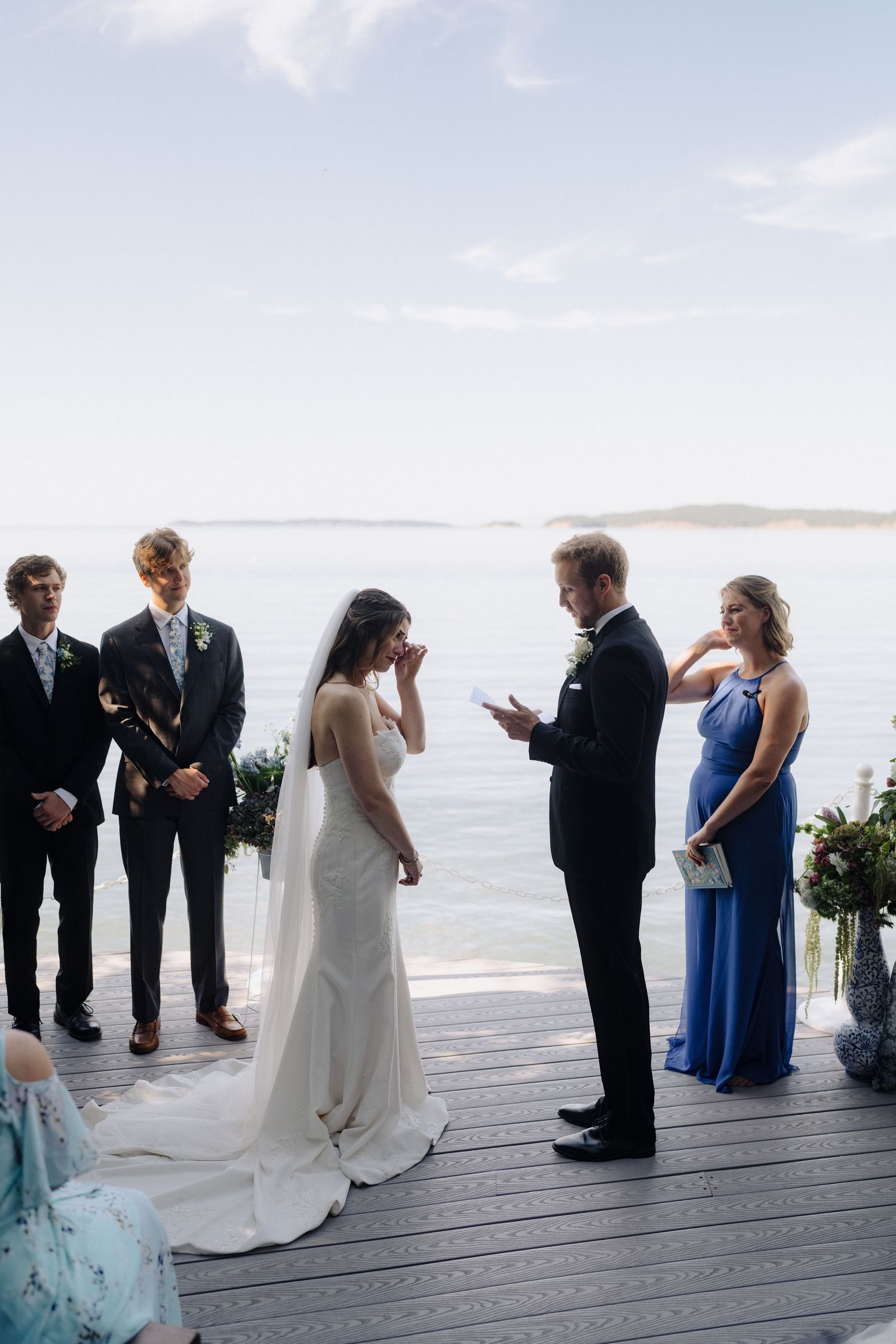 Wedding ceremony participants stand on a wooden boardwalk with a beautiful ocean view in the background.