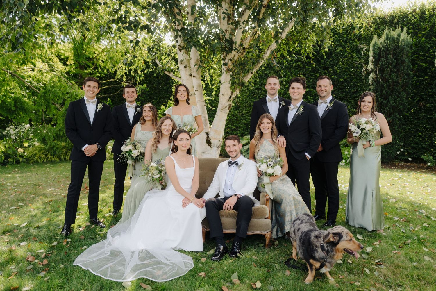 Wedding party poses together on lawn with bridesmaids in sage dresses and groomsmen in black tuxedos.
