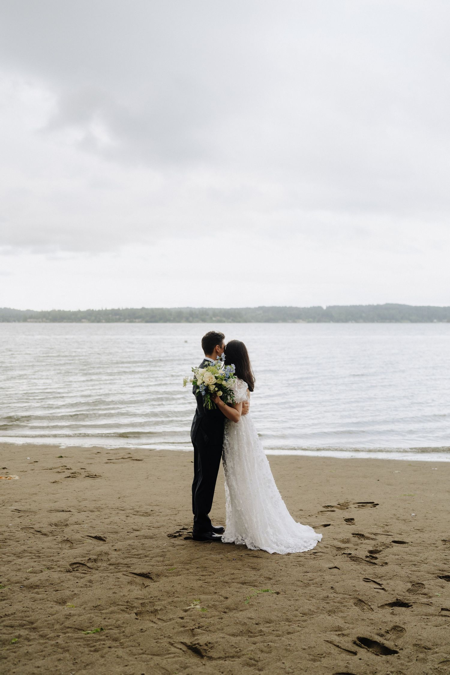 Wedding couple embracing on a misty beach with waves and coastline in the background.
