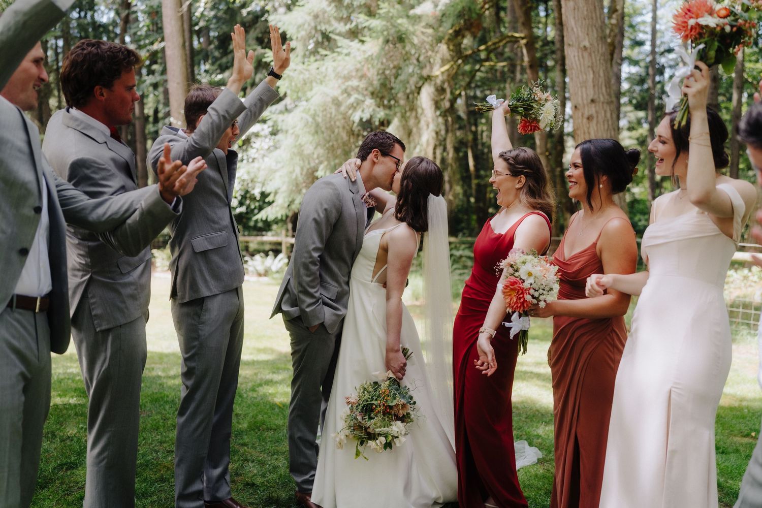 Wedding party celebrates joyfully outdoors with raised hands and bouquets against a backdrop of trees.