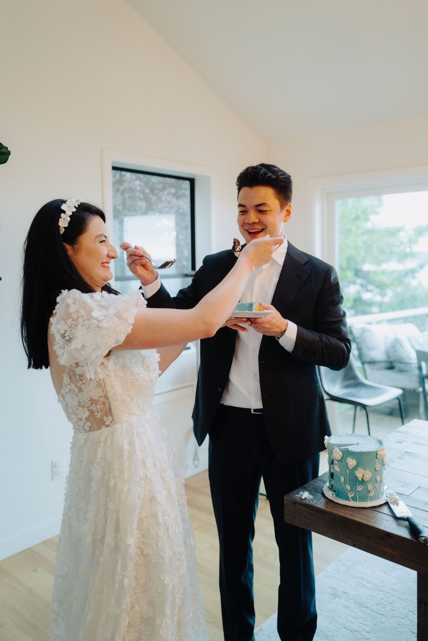 A couple playfully feeding each other wedding cake in a modern white room.