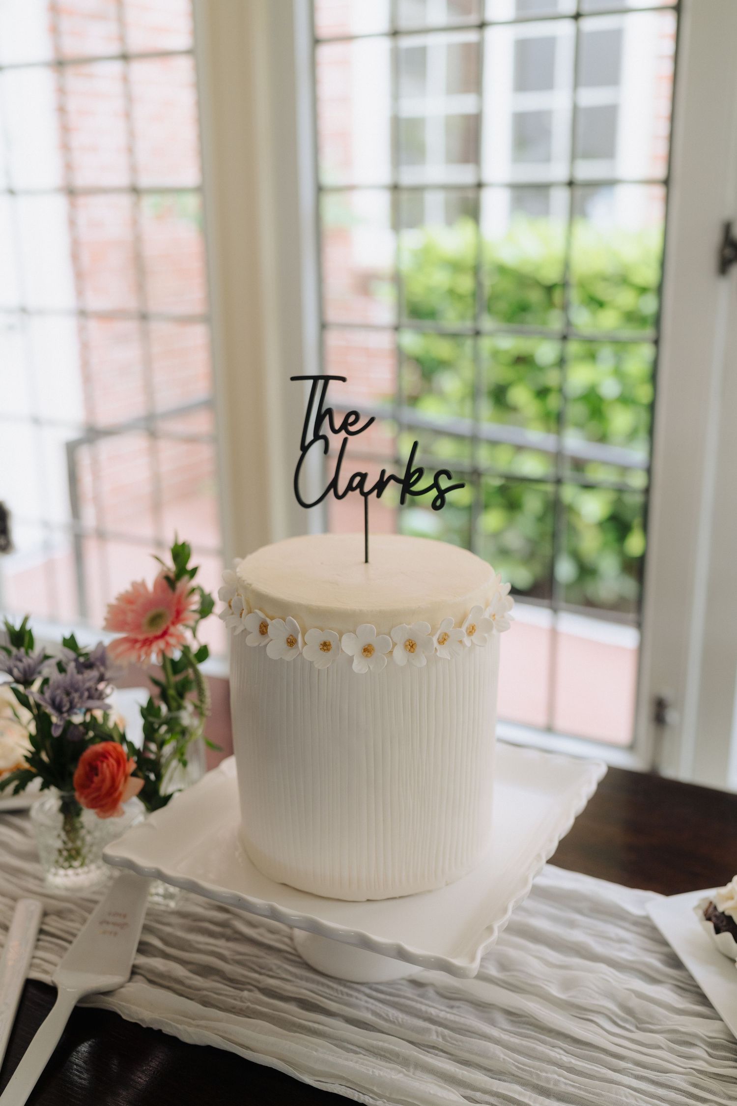 White wedding cakes decorated with fresh flowers displayed on a table by sunny windows.