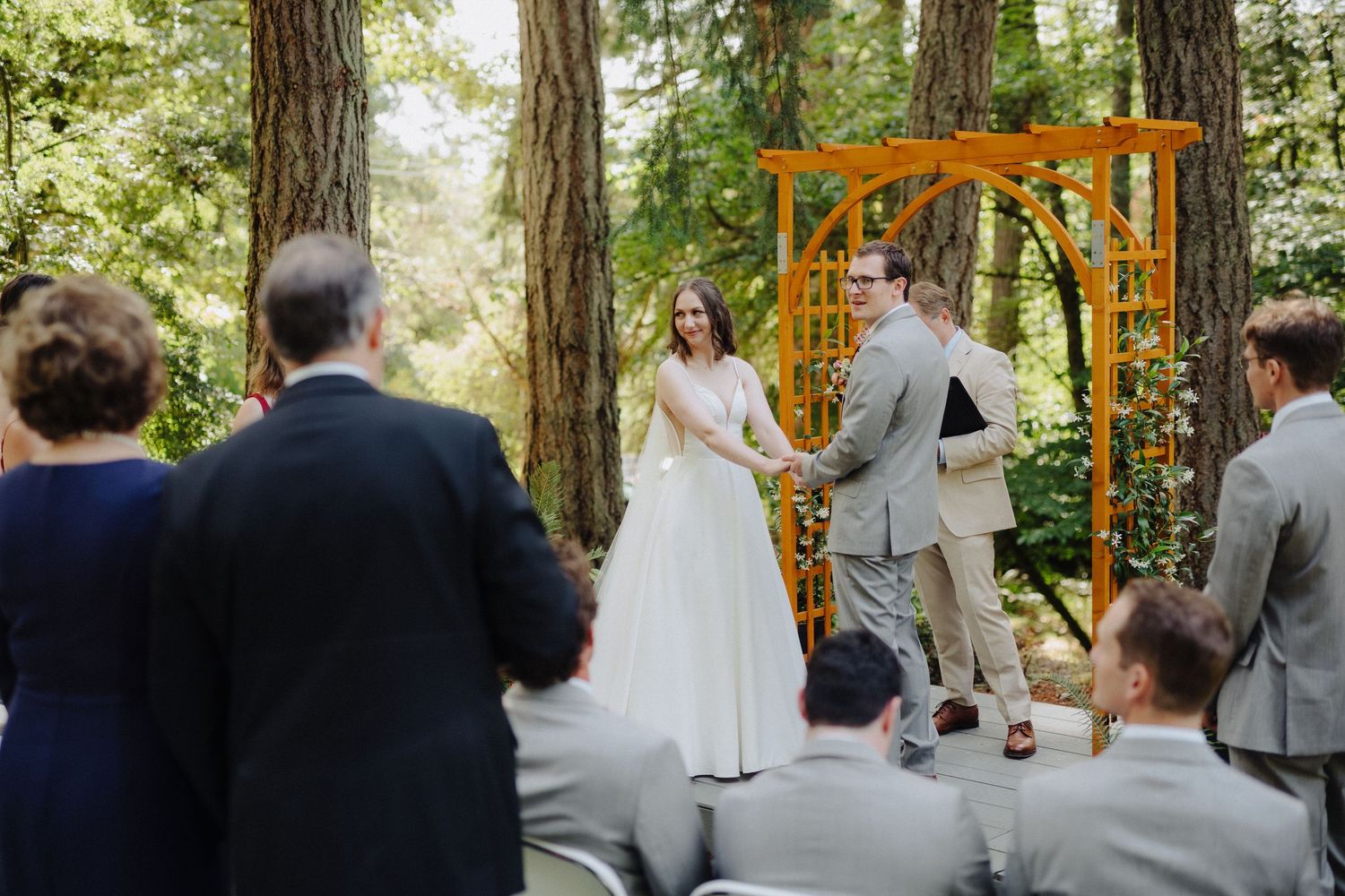 Wedding ceremony taking place in a serene forest setting with an orange arch and guests seated on wooden benches.