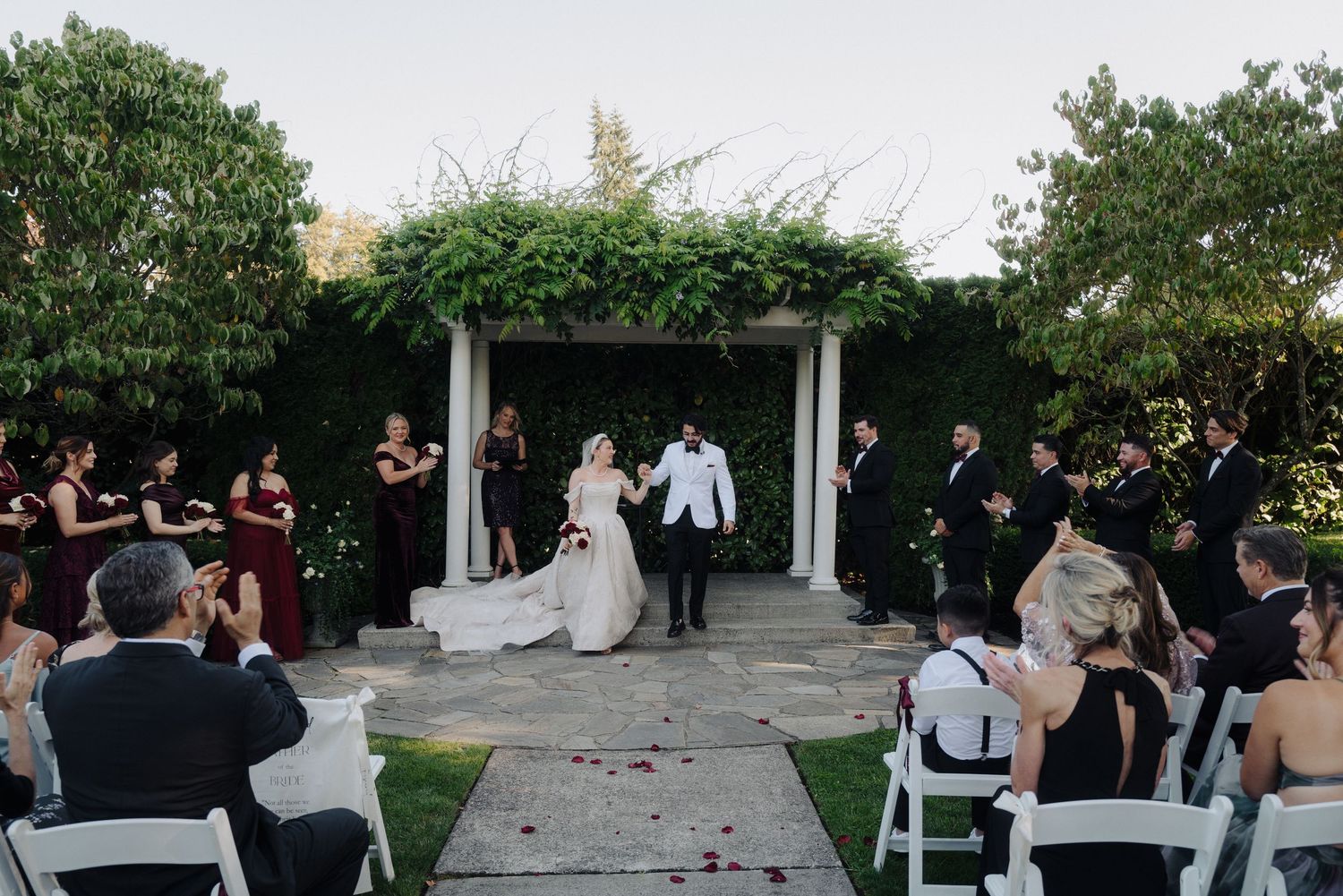 Panoramic view of outdoor wedding ceremony with guests seated in white chairs among garden greenery.