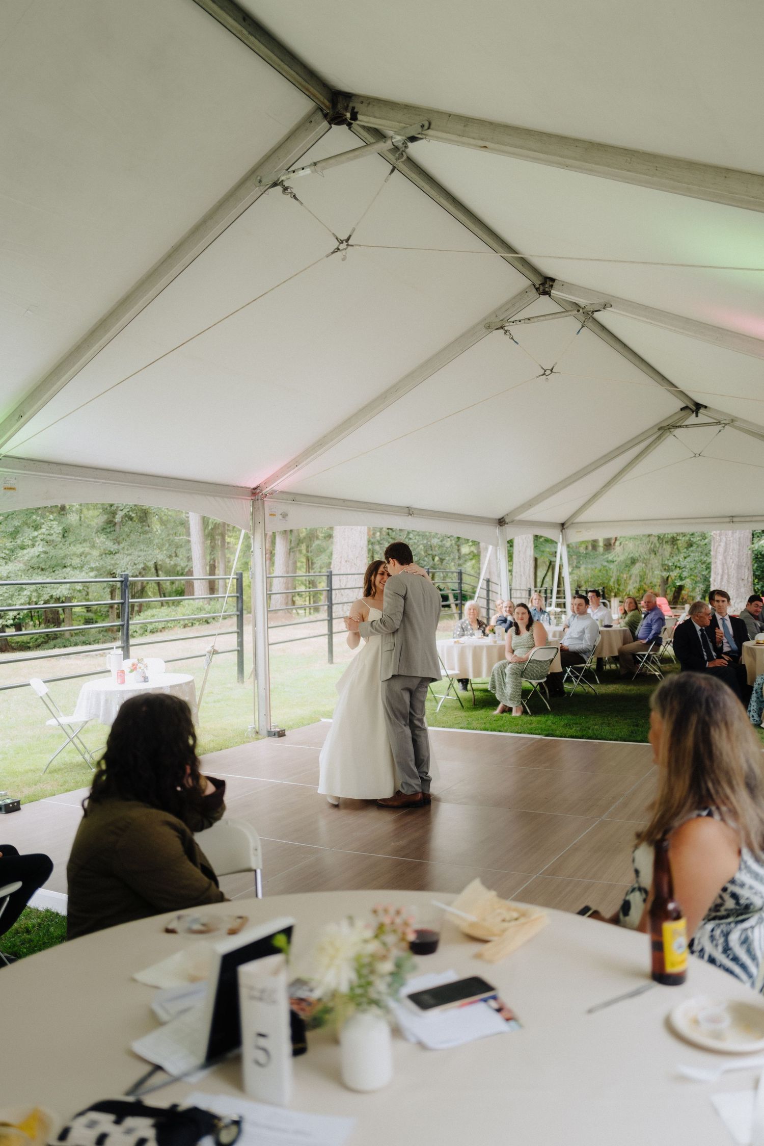 A panoramic view of a white wedding tent reception space with guests seated at tables and a wooden dance floor.