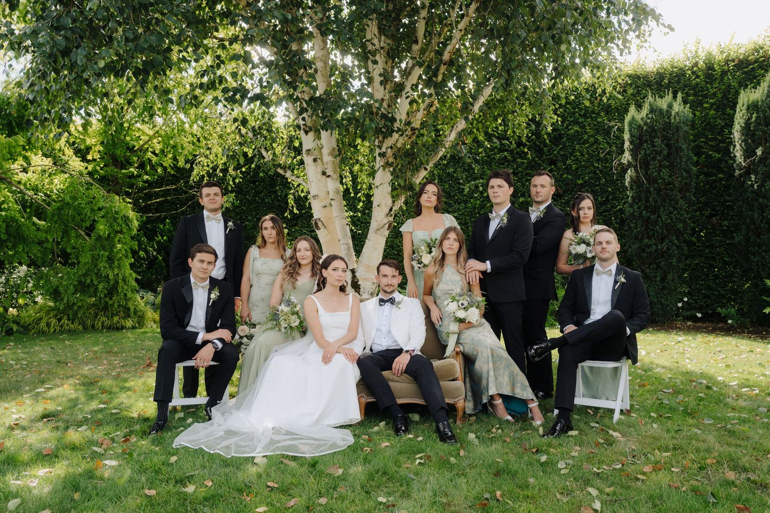Wedding party poses for a formal group photo on a lawn with trees and hedges in the background.