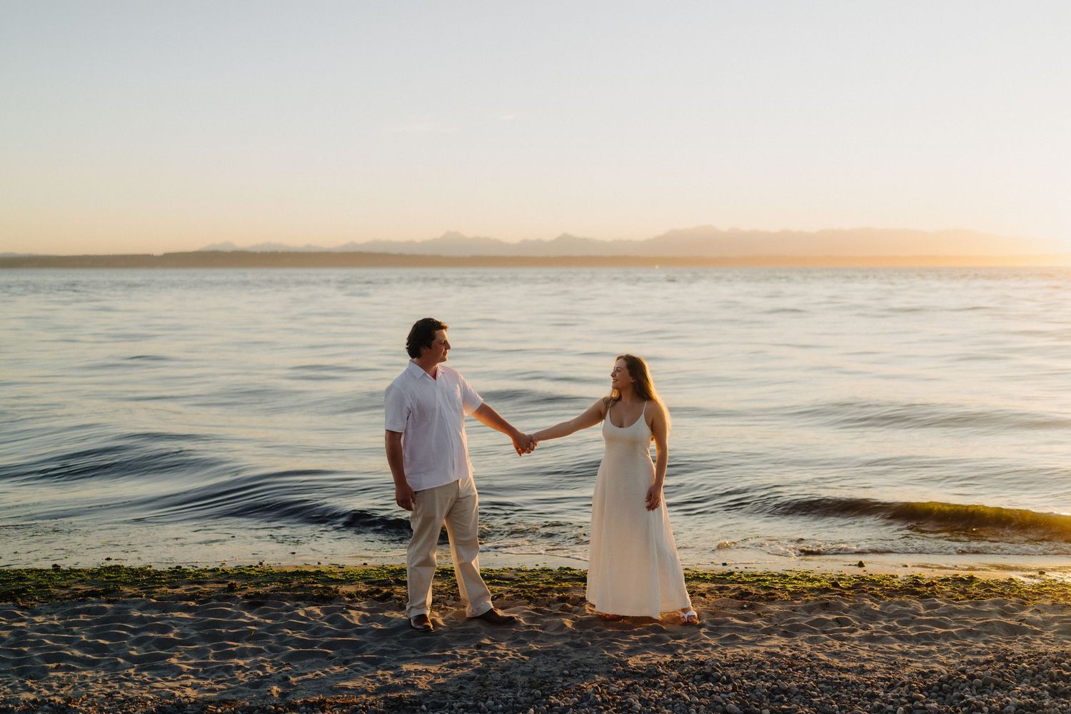 A couple in white outfits sits together on a rocky beach during a beautiful sunset by the ocean.