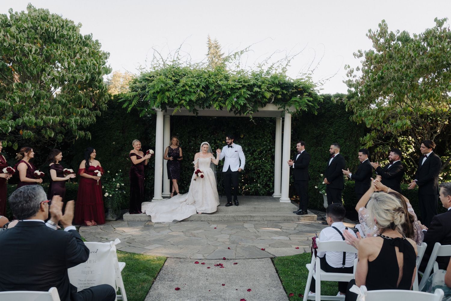 Wedding ceremony taking place under a white pergola with ivy backdrop and seated guests.