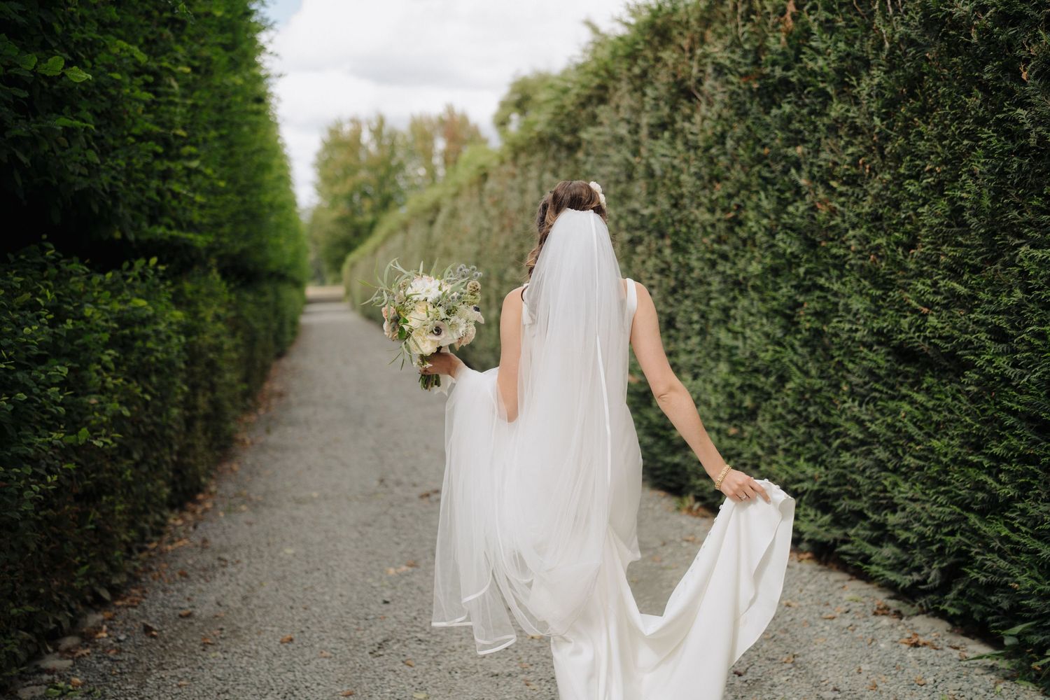 Bride in flowing white dress and veil walks down garden path holding bouquet.