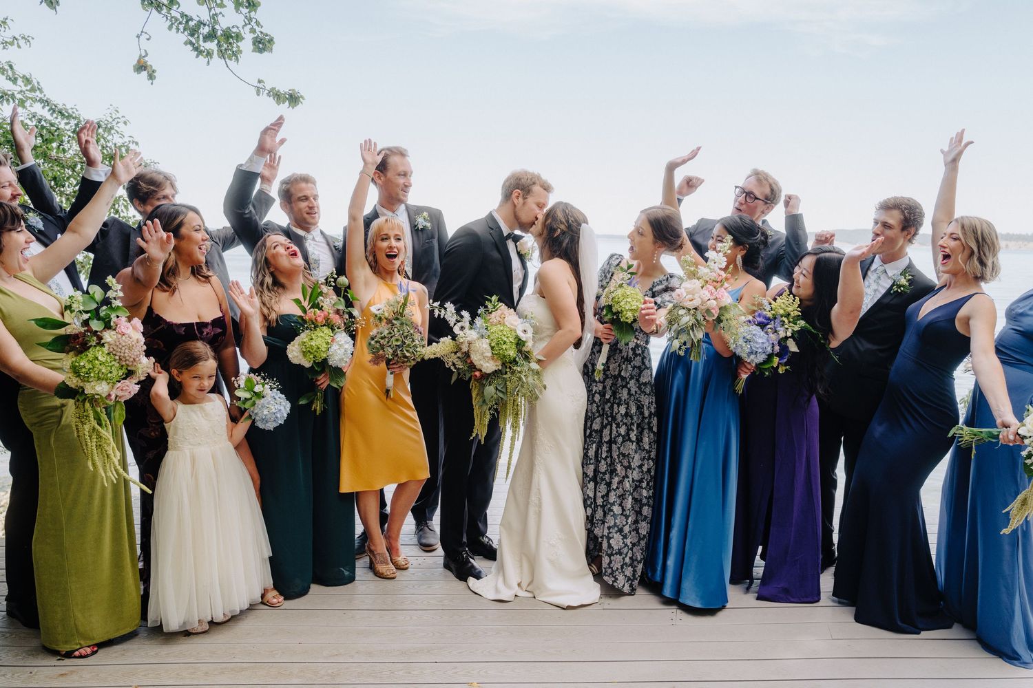 Large wedding party in colorful attire poses together on an outdoor deck with water views.