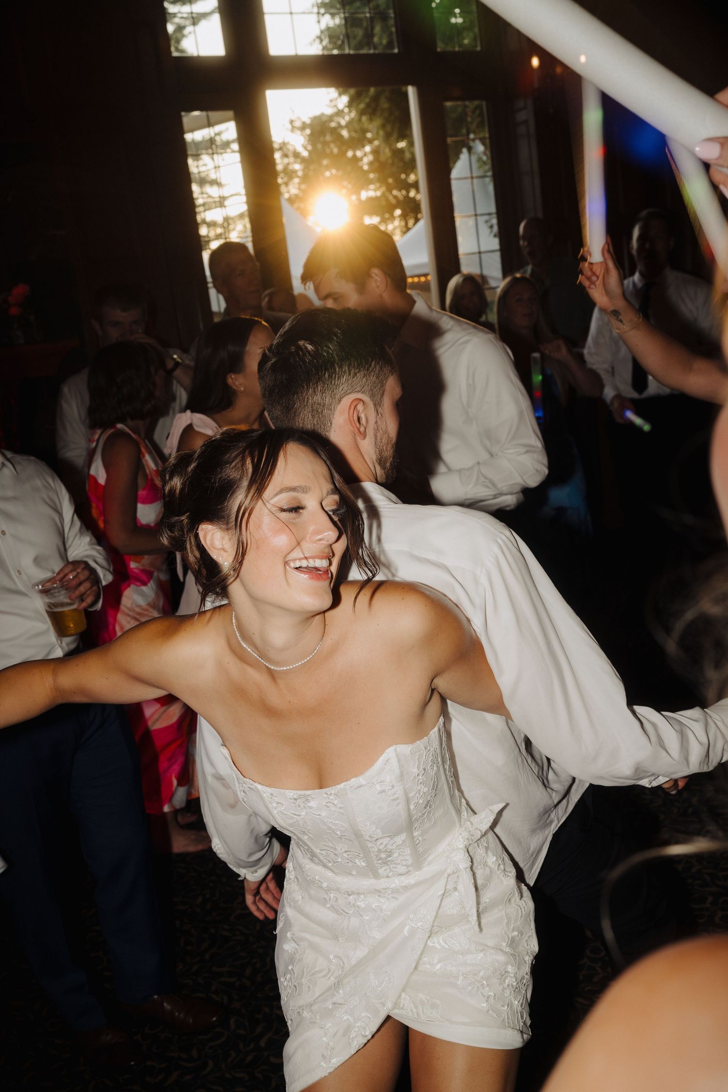 A joyful dance scene at a wedding reception with people celebrating as the sun sets through windows.