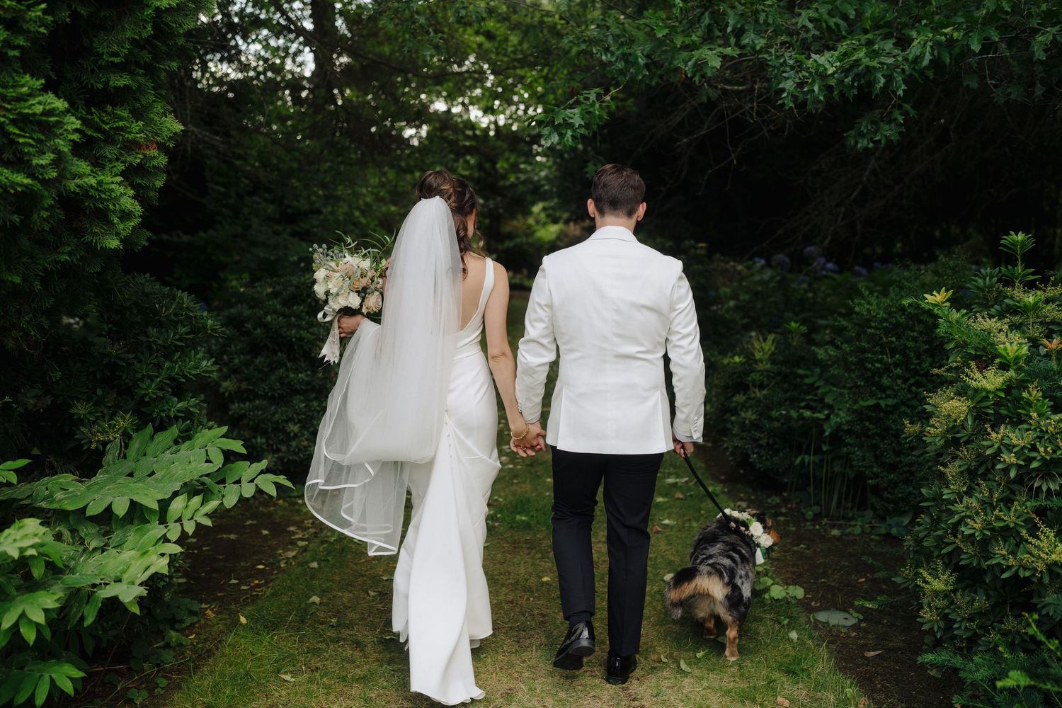 Wedding party walking through a garden pathway with trees and greenery in the background.