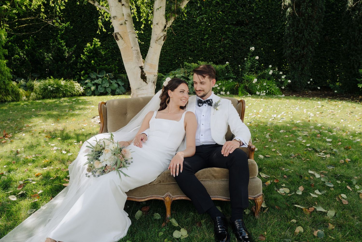 Newlyweds share a romantic moment on a bench in a lush garden setting during their wedding day.