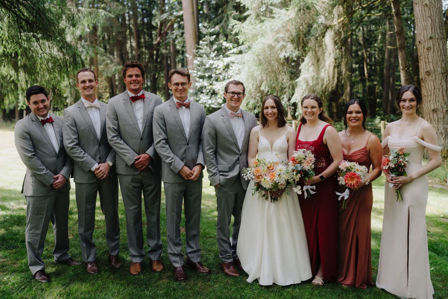 Wedding party in burgundy and gray attire posing together in a forest setting.